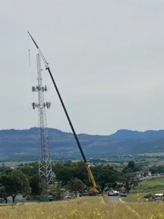 Long-boom crane by Jacked Co. reaching a high-altitude cellular tower for telecommunications infrastructure maintenance.