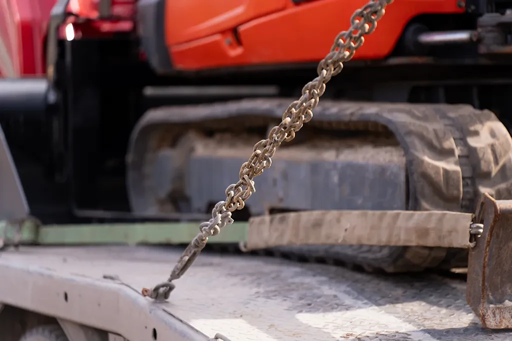 Close-up of heavy equipment secured to a flatbed trailer with high-tension steel chains and transport straps.