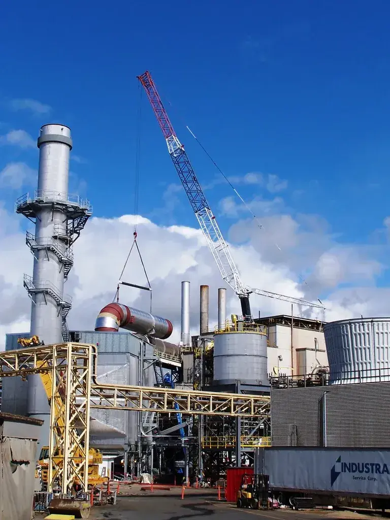 Lattice boom crane performing a heavy lift of industrial ductwork at a processing mill or power plant.