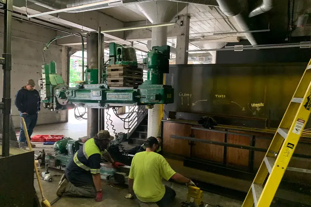 Jacked Co. rigging team carefully decommissioning and moving a large green radial drill press in a factory.