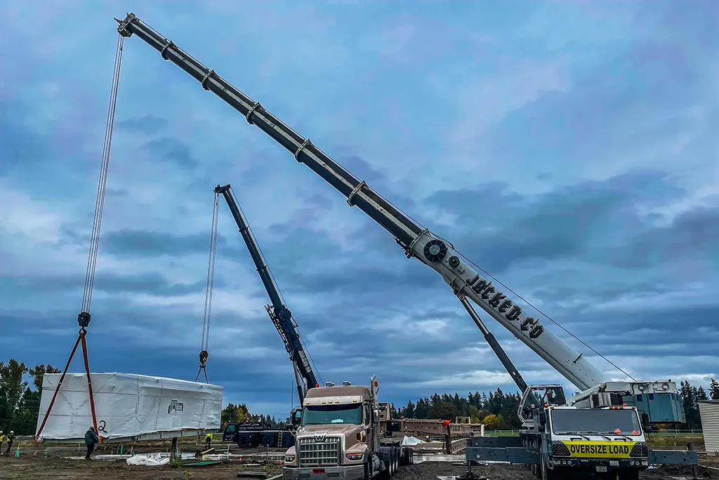 Tandem lift by Jacked Co. cranes moving a large, wrapped industrial unit at a construction site.