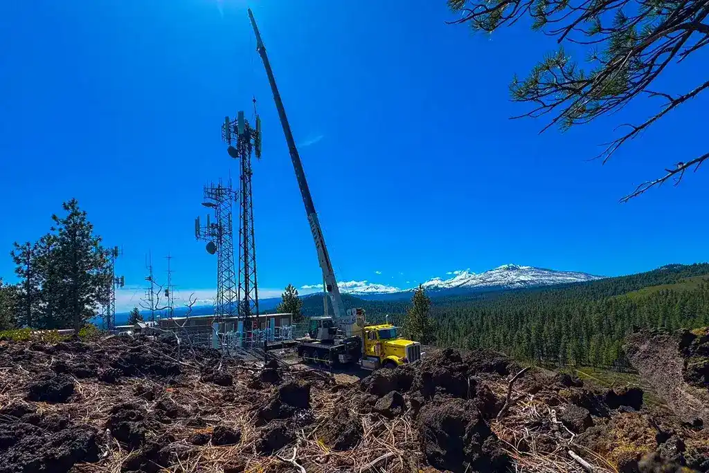 Mobile crane by Jacked Co. servicing a mountain-top telecommunications site with a view of snow-capped peaks.