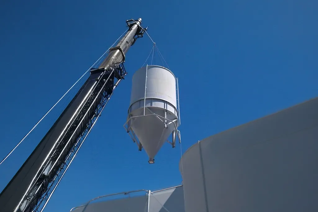 Vertical shot of a Jacked Co. crane performing a high lift of a large white industrial silo against a clear blue sky.