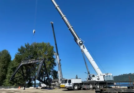 Dual mobile cranes from Jacked Co. performing a tandem lift of a massive steel bridge section near a waterway.