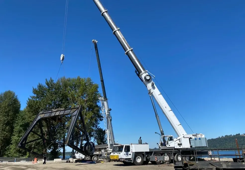Dual mobile cranes from Jacked Co. performing a tandem lift of a massive steel bridge section near a waterway.