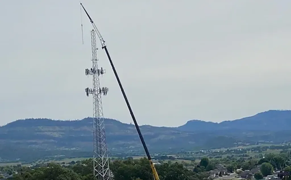 Long-boom crane reaching the top of a telecommunications lattice tower for antenna installation or repair.