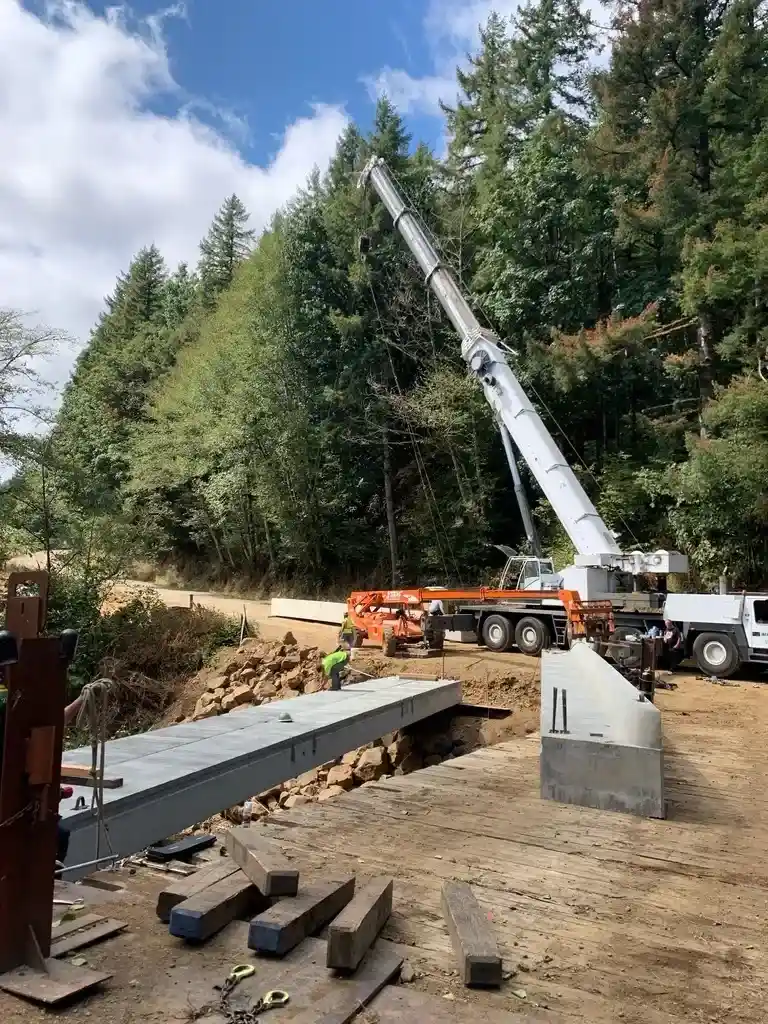 Mobile crane from Jacked Co. installing concrete bridge girders for a rural infrastructure project in a forested area.