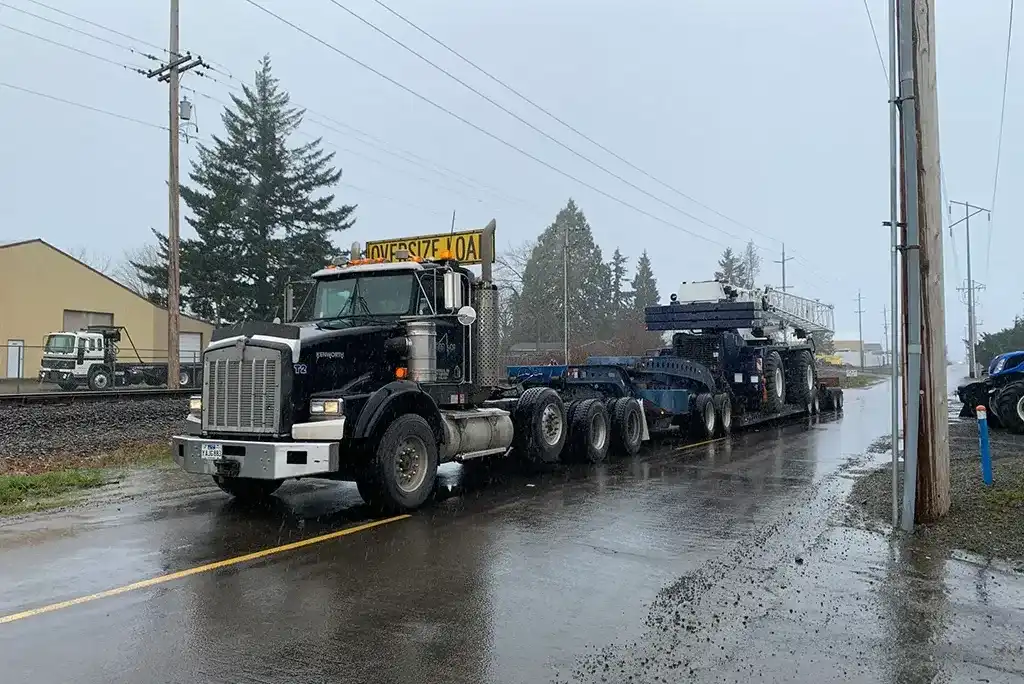 Kenworth heavy-haul truck from Jacked Co. transporting a massive mobile crane chassis on a multi-axle trailer in the rain.