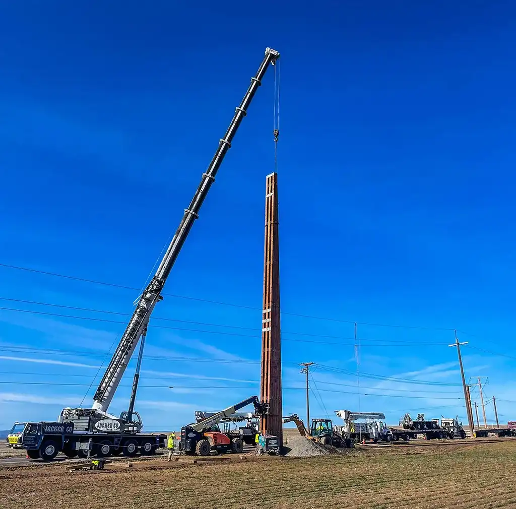 A white Jacked Co mobile crane lifting a tall vertical wooden column on an open construction site, surrounded by heavy equipment and utility vehicles.