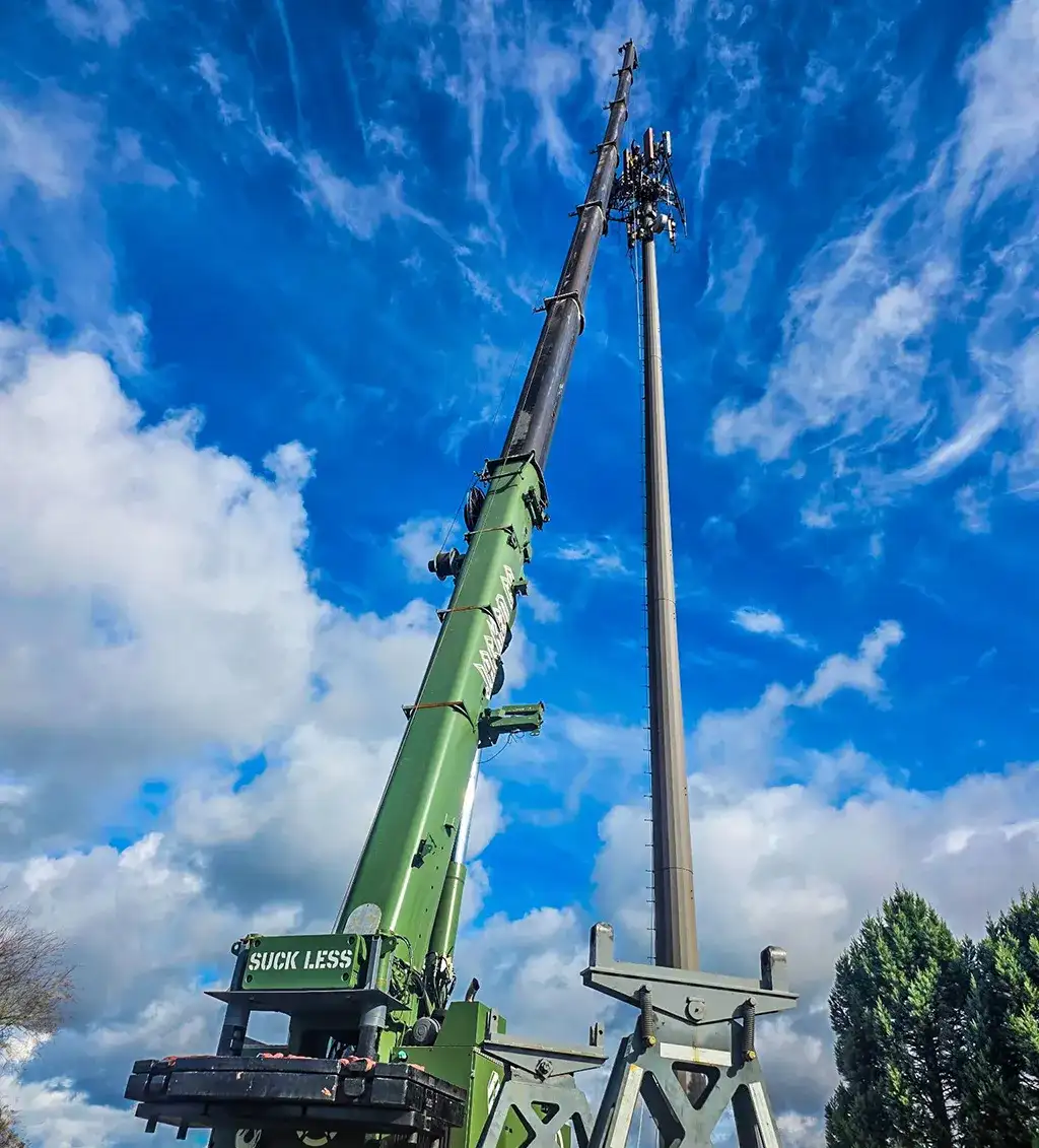 A green Jacked Co crane with its boom fully extended upwards, performing maintenance work next to a tall telecommunications cell tower against a partly cloudy sky.