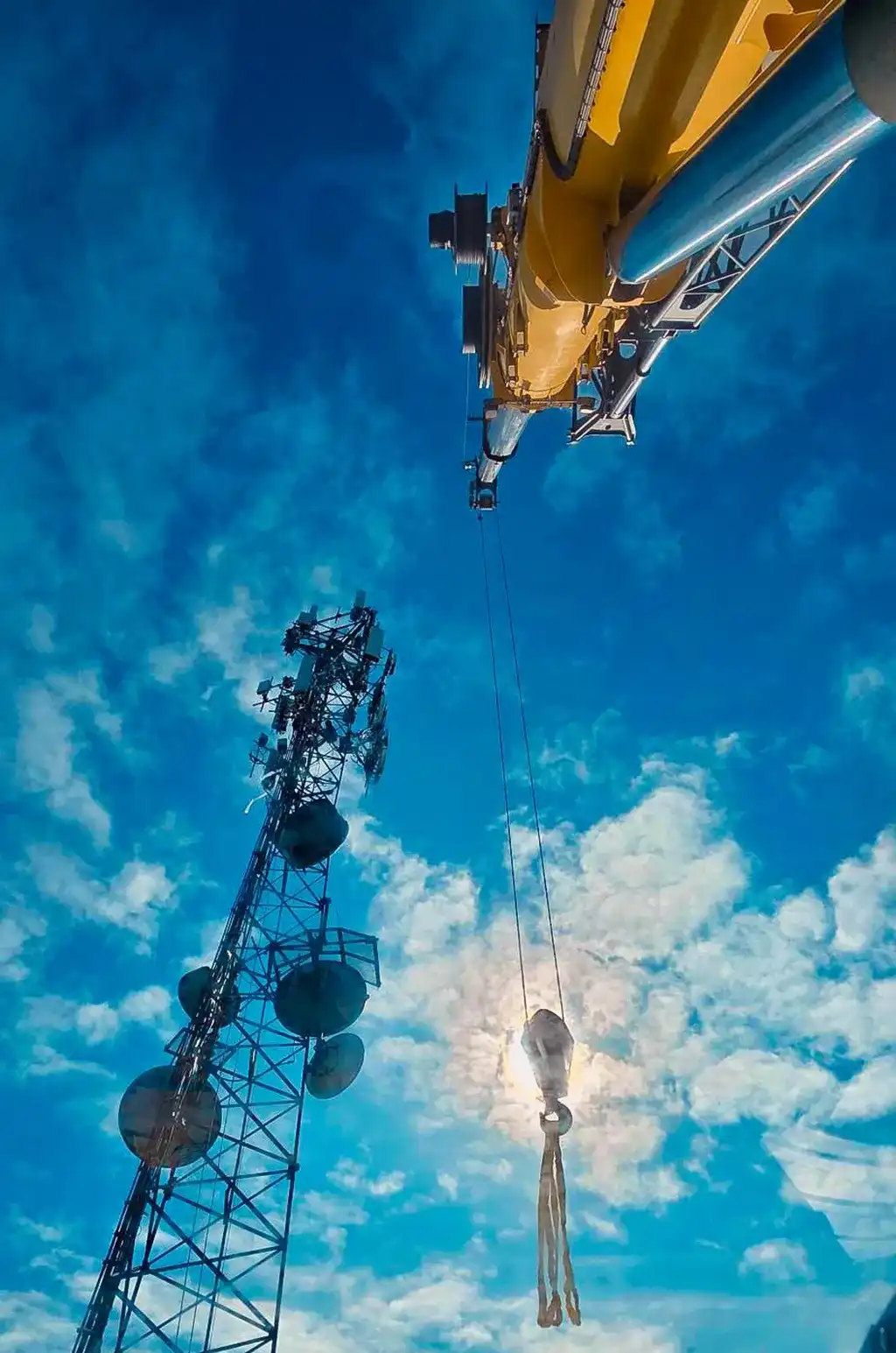 A dynamic low-angle shot looking straight up at a yellow crane boom and its hanging hook, positioned next to a tall radio tower with the sun backlighting the rigging.