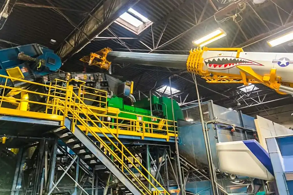 An indoor shot of a yellow Jacked Co crane with a shark mouth design precision-lifting a component over complex green and blue industrial machinery inside a factory.