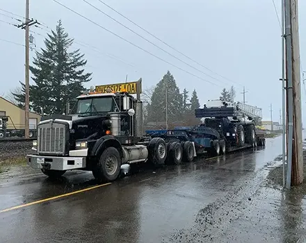 Kenworth heavy-haul truck from Jacked Co. transporting a massive mobile crane chassis on a multi-axle trailer in the rain.