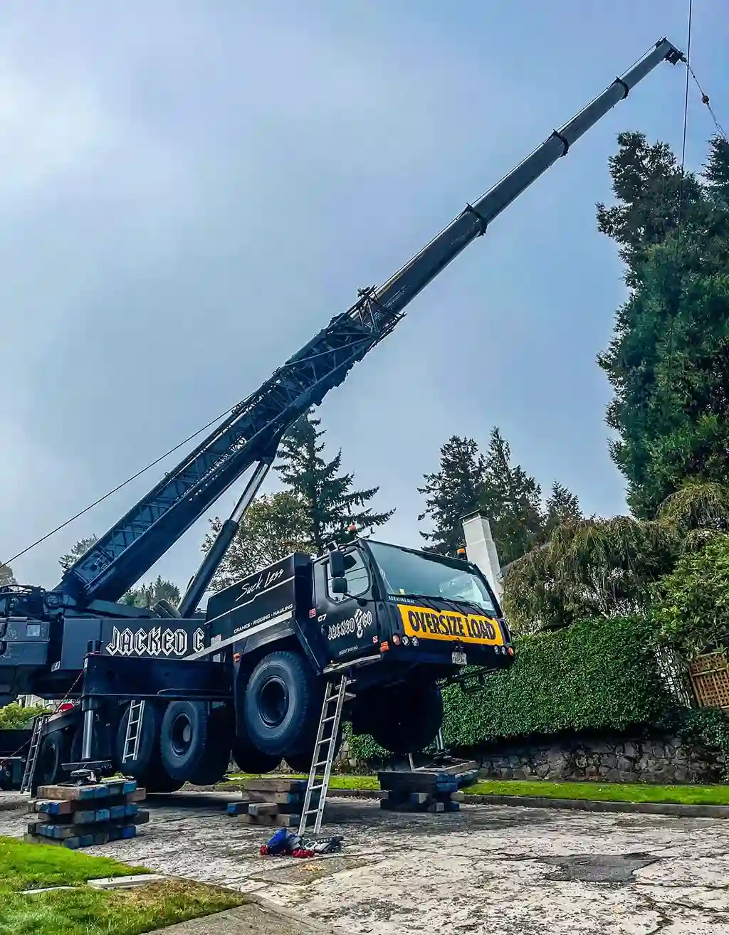 A heavy-duty black Jacked Co crane displaying an "OVERSIZE LOAD" banner, stabilized on extensive wooden cribbing and outriggers, extending its boom over tall pine trees in a residential neighborhood.