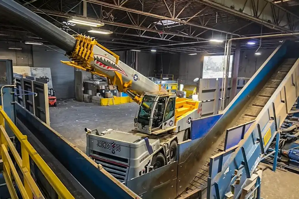 A specialized yellow and white Jacked Co crane featuring a shark mouth decal operating indoors, maneuvering its boom around large conveyors inside an industrial manufacturing facility.