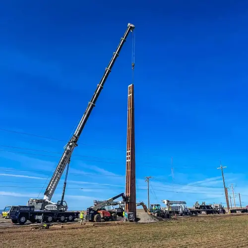 A large Jacked Co. mobile crane hoisting a tall, vertical wooden transmission pole into position at a construction site under a clear blue sky