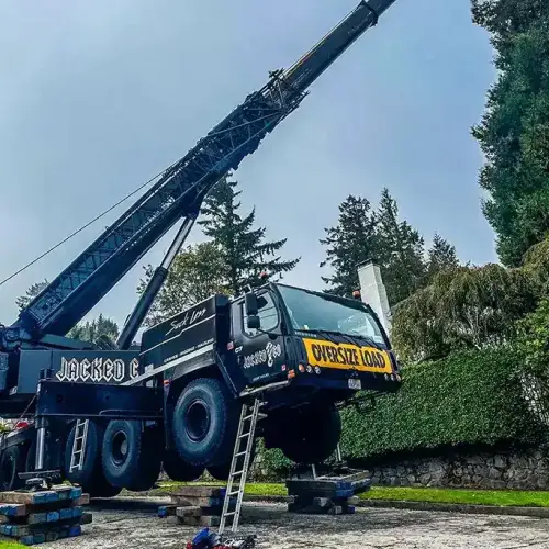 A Jacked Co crane stabilized on wooden cribbing and outriggers, extending its boom over tall pine trees in a residential neighborhood.